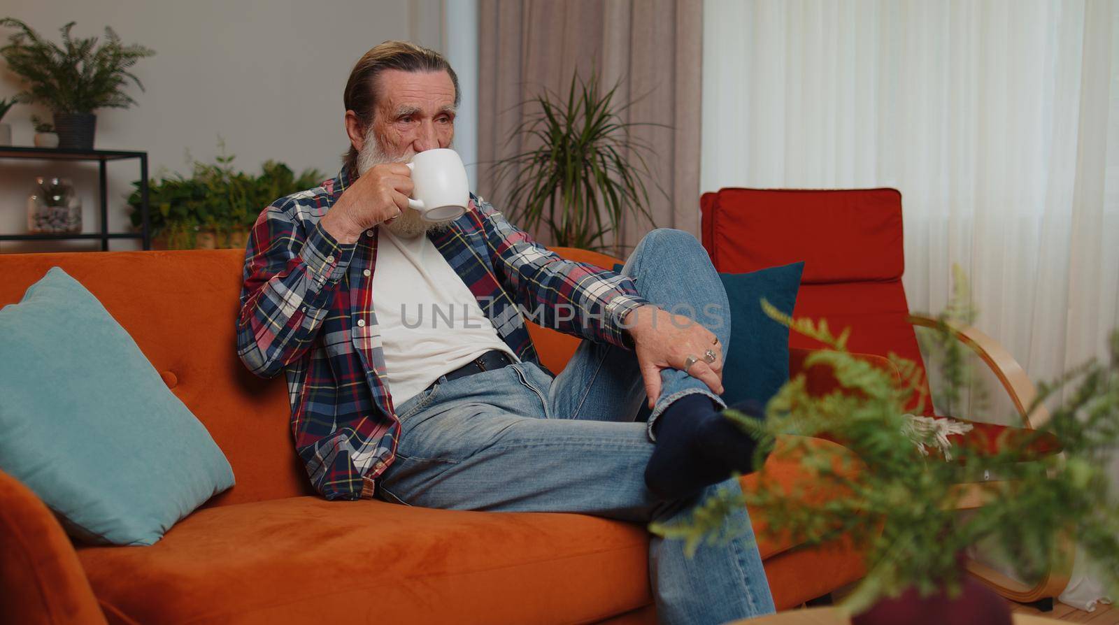 Attractive smiling senior old grandfather man drinking a cup of warm coffee or herbal tea sitting at home living room couch in the morning. Elderly retired grandpa pensioner enjoying comfortable relax