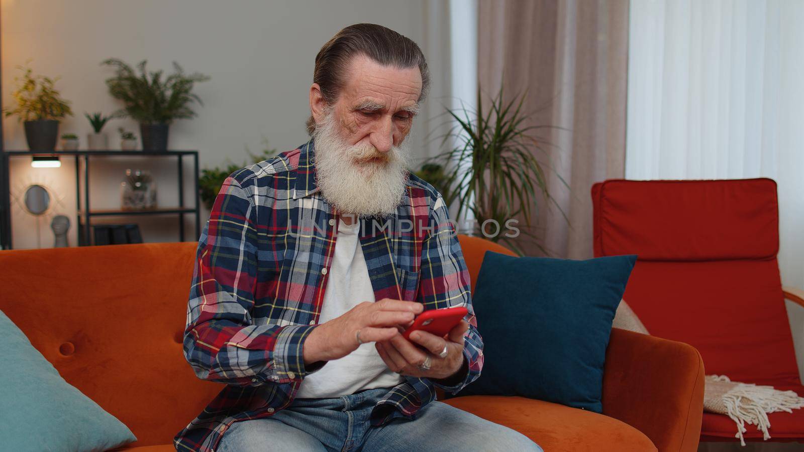 Senior old grandfather sitting on sofa uses mobile phone smile at modern home apartment. Elderly man texting share messages content on smartphone social media applications online, watching relax movie