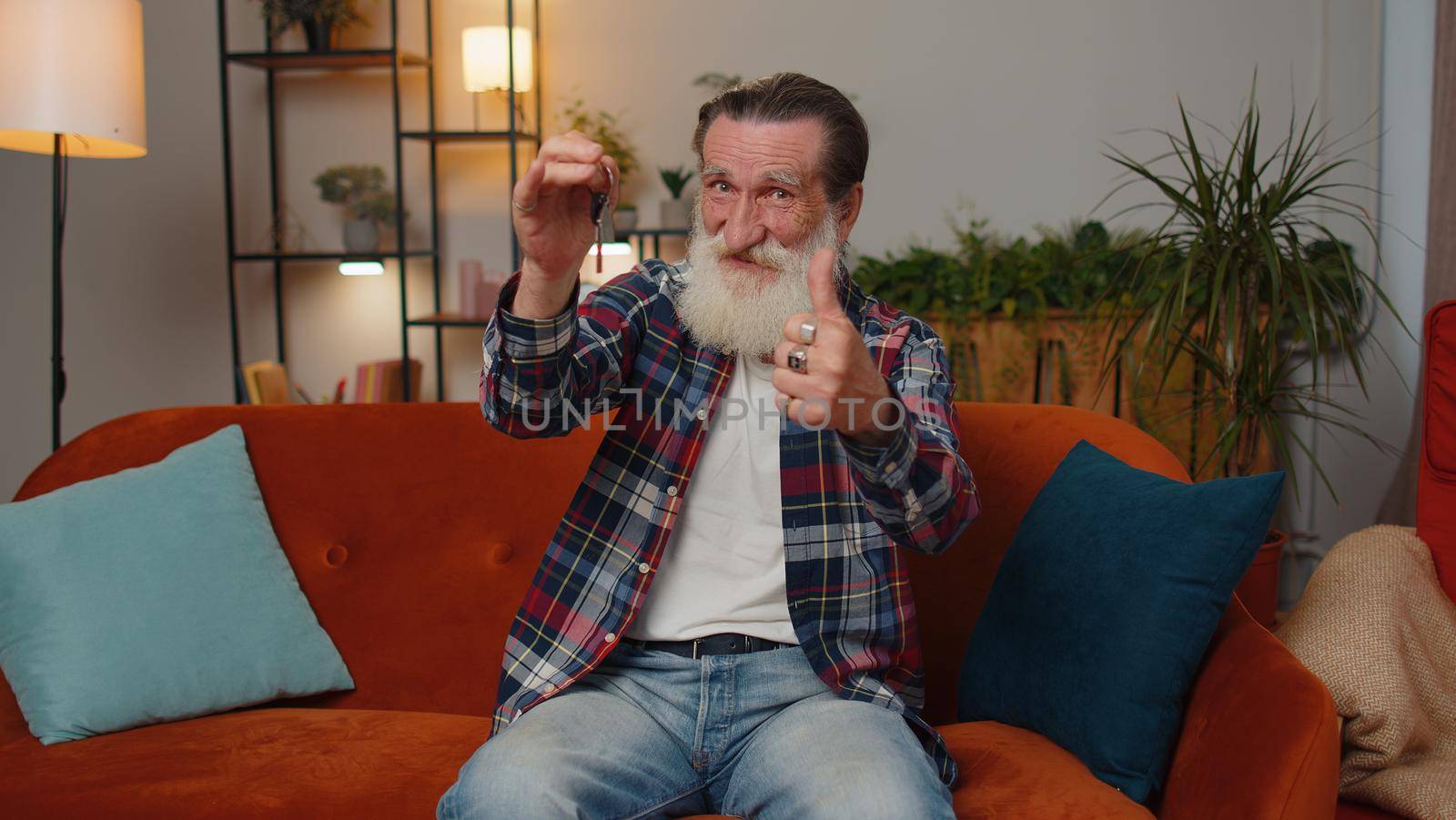 Senior old grandfather real estate agent lifting hand showing the keys of new home house apartment, buying or renting property, mortgage loan at modern home apartment indoors. Man in room sits on sofa
