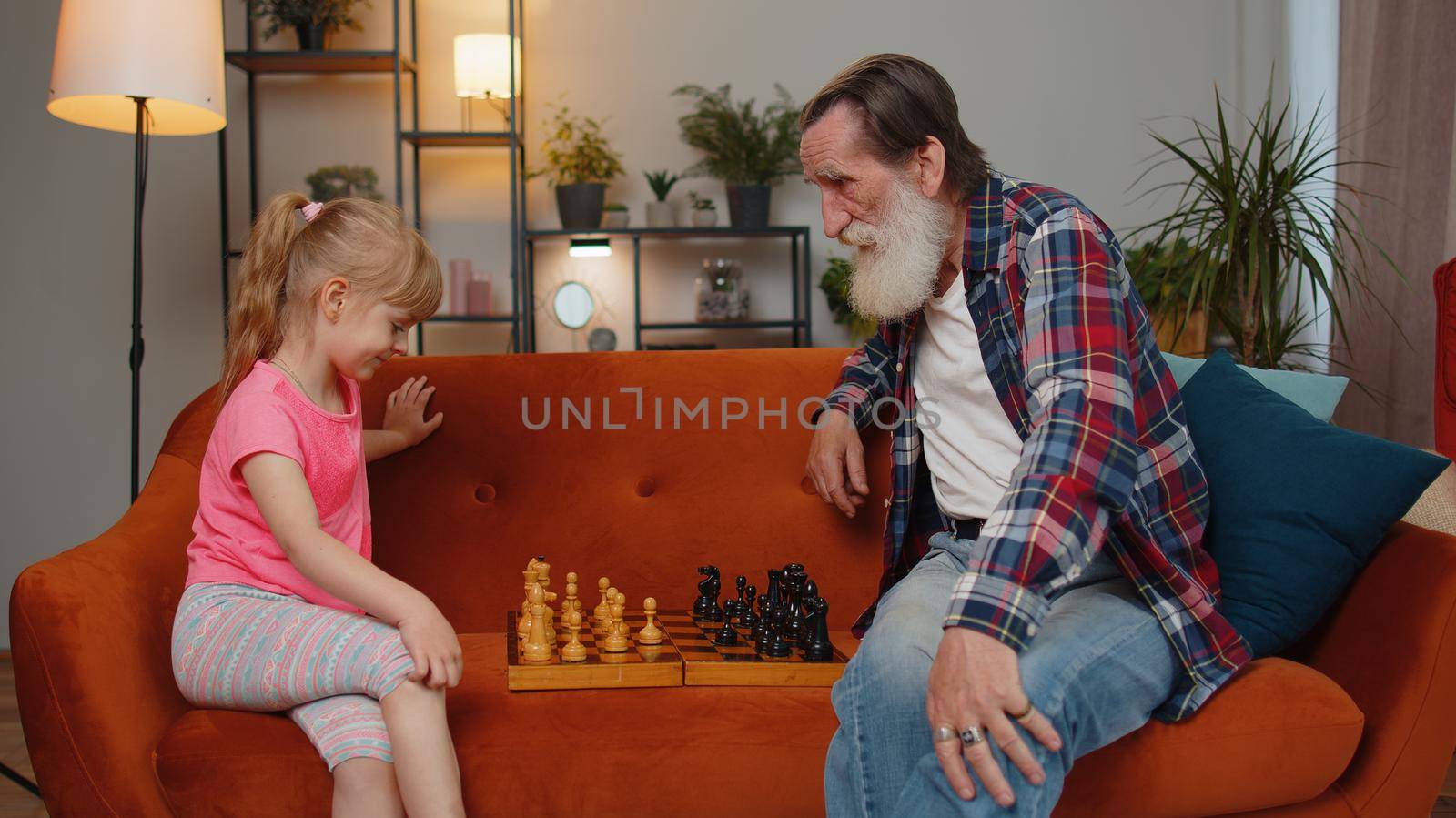 Smiling senior grandfather man playing chess with teen granddaughter at home living room. Retired grandparent enjoying play board game, resting on couch with child girl kid. Family domestic activity