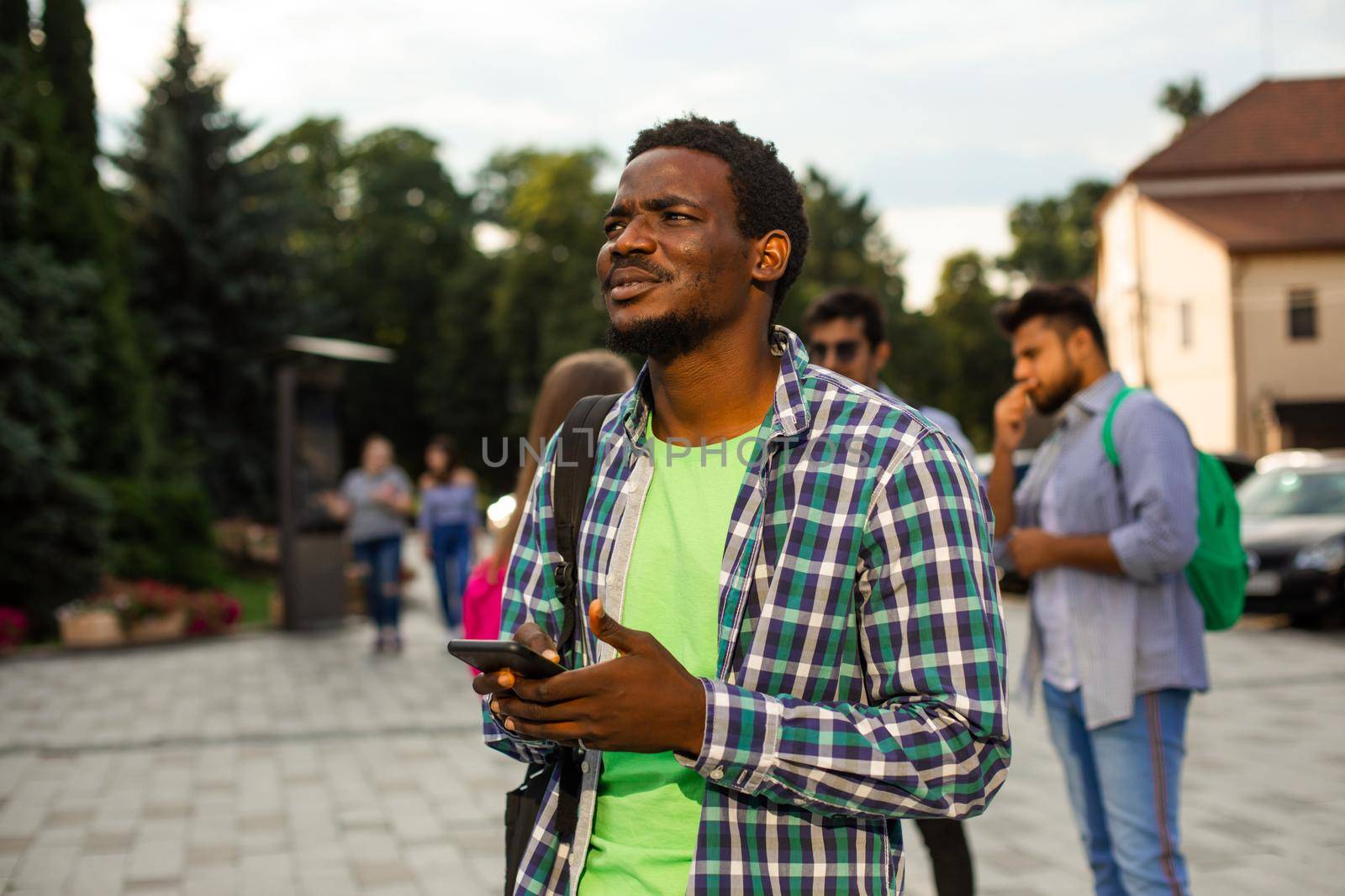 The young african student with a bag outdoors is holding a phone. The man in a shirt is looking for road to home with help navigation