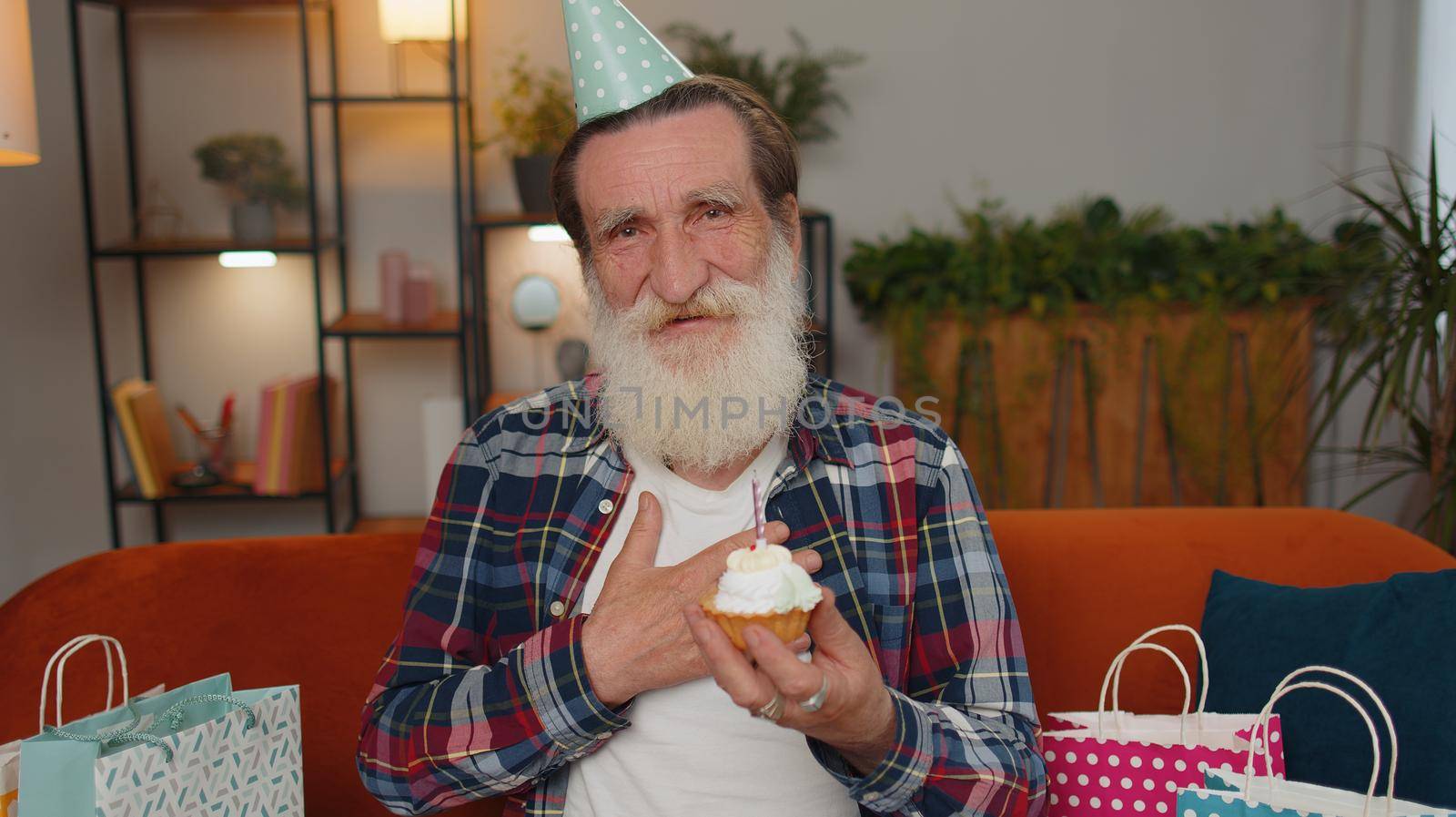Happy senior grandfather wears festive birthday hat hold cupcake makes wish joyful congratulating blowing burning candle on cake. Mature man celebrating anniversary party on couch with gifts at home