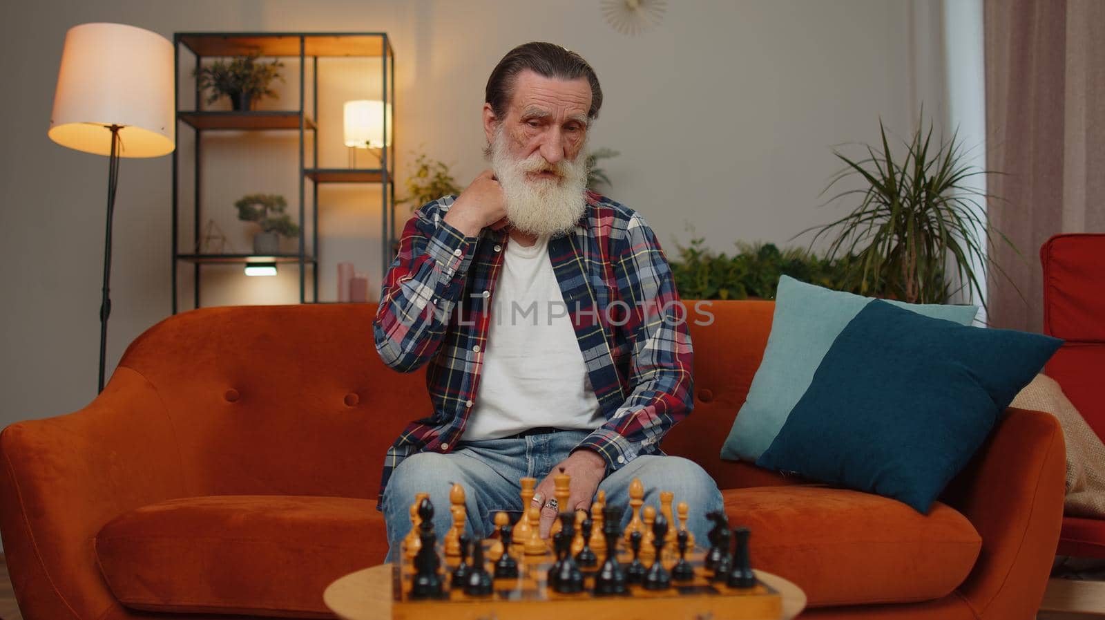 Focused senior grandfather man chessman sitting at home room couch learning playing chess board game alone, educational leisure domestic activity, strategic skills. Elderly retired grandpa pensioner
