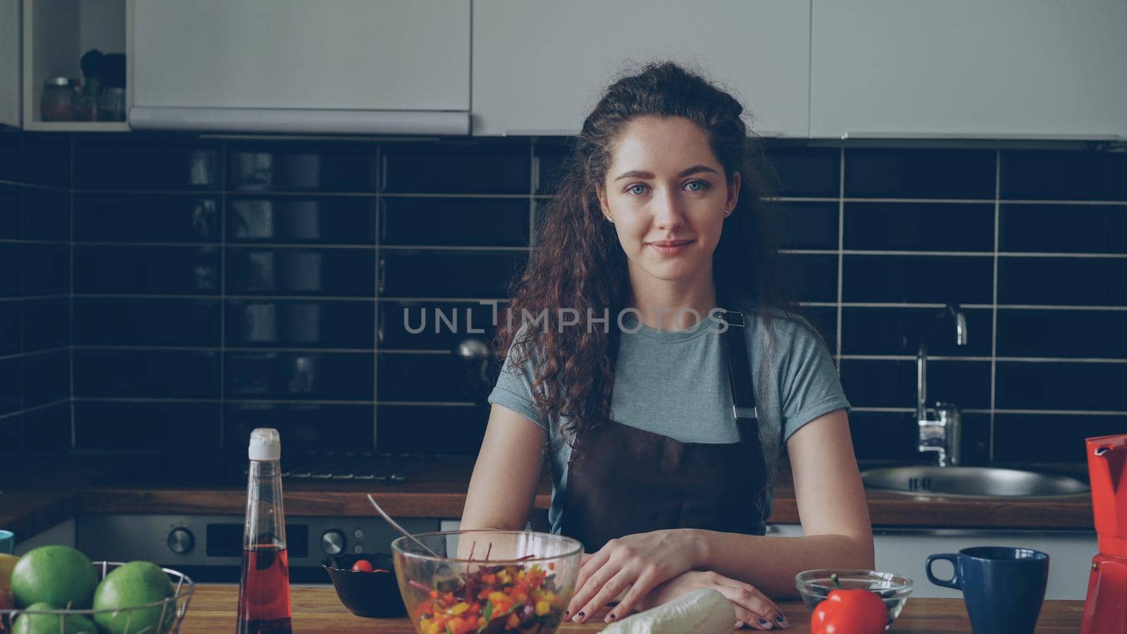 Portrait of young caucasian executive chef woman in apron sitting at table in modern lighty spacious kitchen, smiling,calm and positive, looking at camera by silverkblack