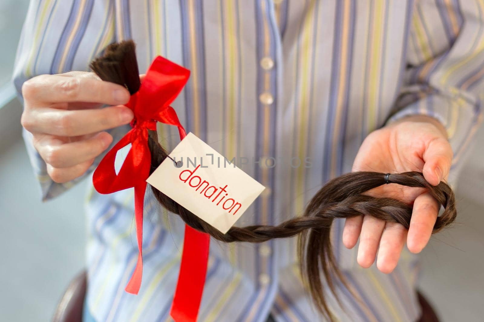 Long brown cut braid with tag and red ribbon bow in woman hands, donation of haircut, female faceless concept