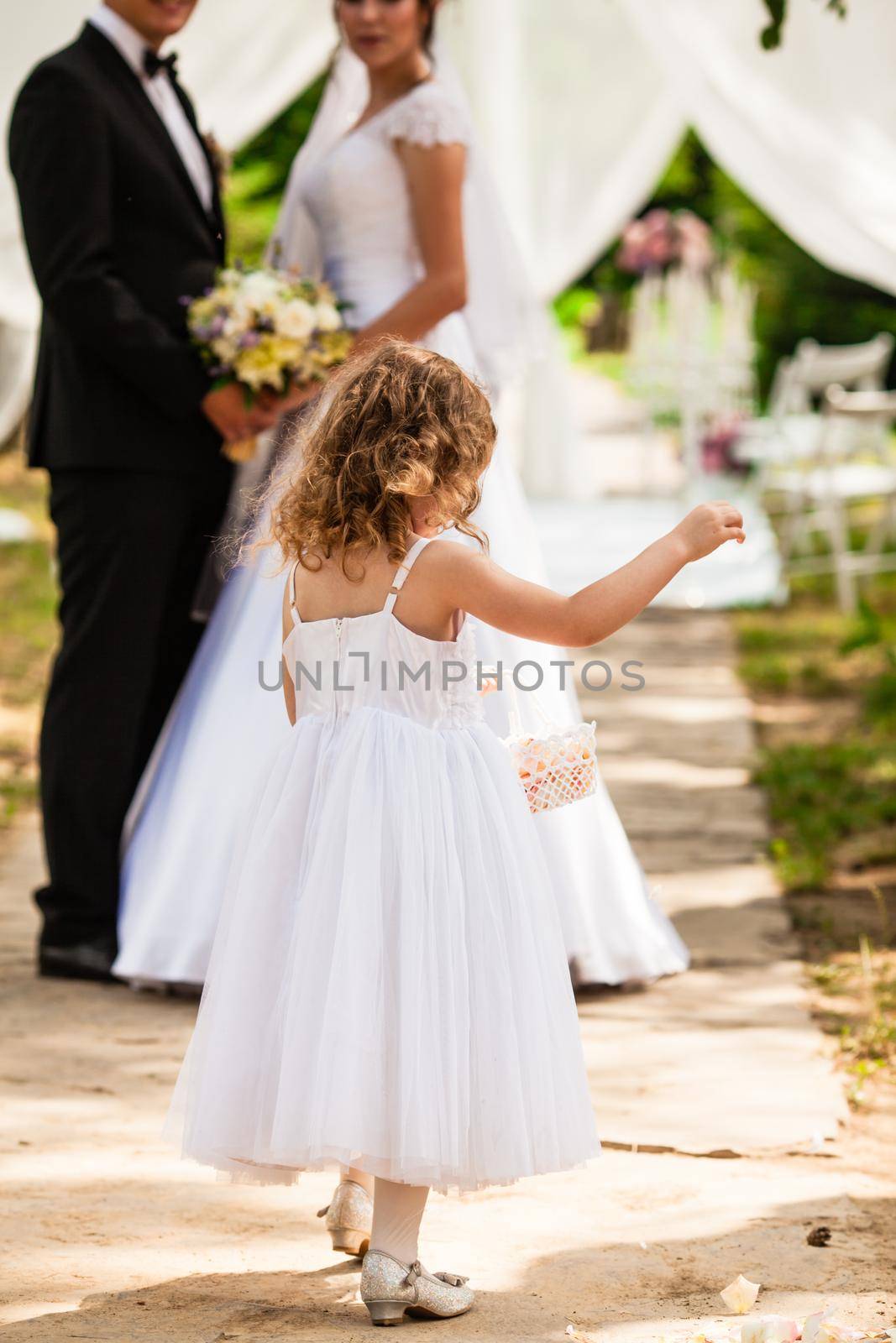 Cute little bridesmaid with a basket in which the petals of roses looks into the camera with wedding couple in background