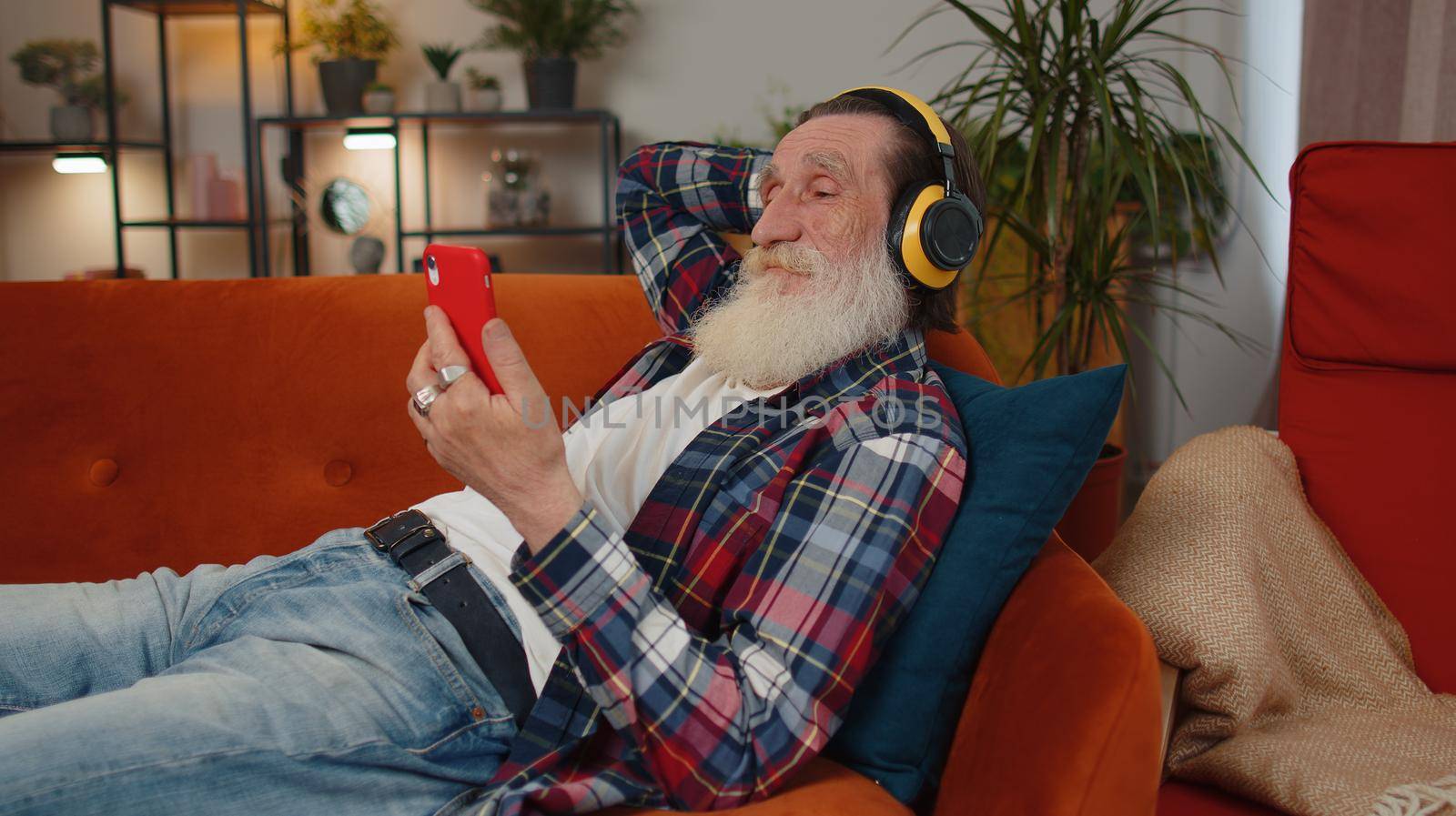 Happy relaxed overjoyed senior man in wireless headphones dancing on couch at home choosing listening favorite energetic disco rock n roll music in smartphone entertaining. People weekend activities