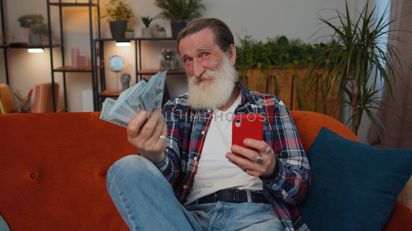 Planning family budget. Smiling senior old grandfather counting money cash, use smartphone calculate domestic bills at home. Joyful man satisfied of income and saves money for planned vacation, gifts