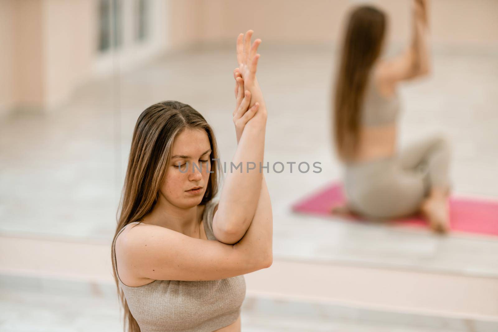 Young woman doing yoga in the gym. A girl with long hair and in a beige tracksuit stands in a cow pose on a pink carpet. A woman performs Gomukhasana. by Matiunina
