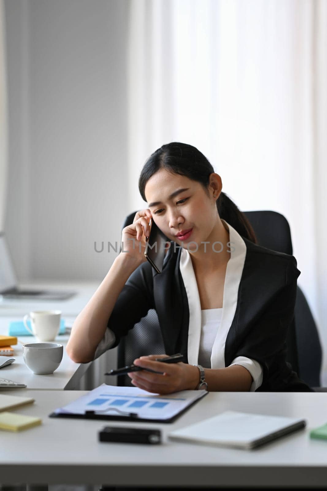 Attractive businesswoman talking on mobile phone while sitting in modern office. by prathanchorruangsak