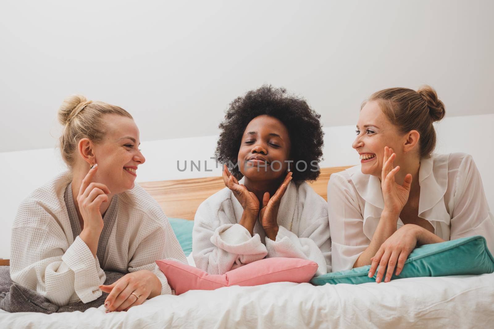 The young women are lying in bathrobes on the bed and communicate. The happy girlfriends are relaxing at the spa hotel