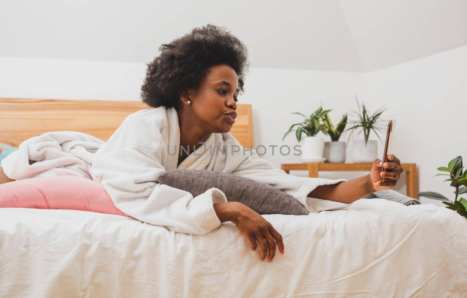 The beautiful woman is lying on the bed in a bathrobe and surfing the internet. The woman with smartphone is resting in a hotel