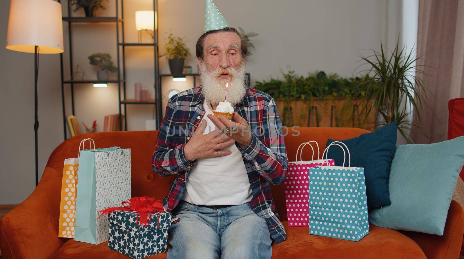 Happy senior grandfather wears festive birthday hat hold cupcake makes wish joyful congratulating blowing burning candle on cake. Elderly man celebrating anniversary party on couch with gifts at home