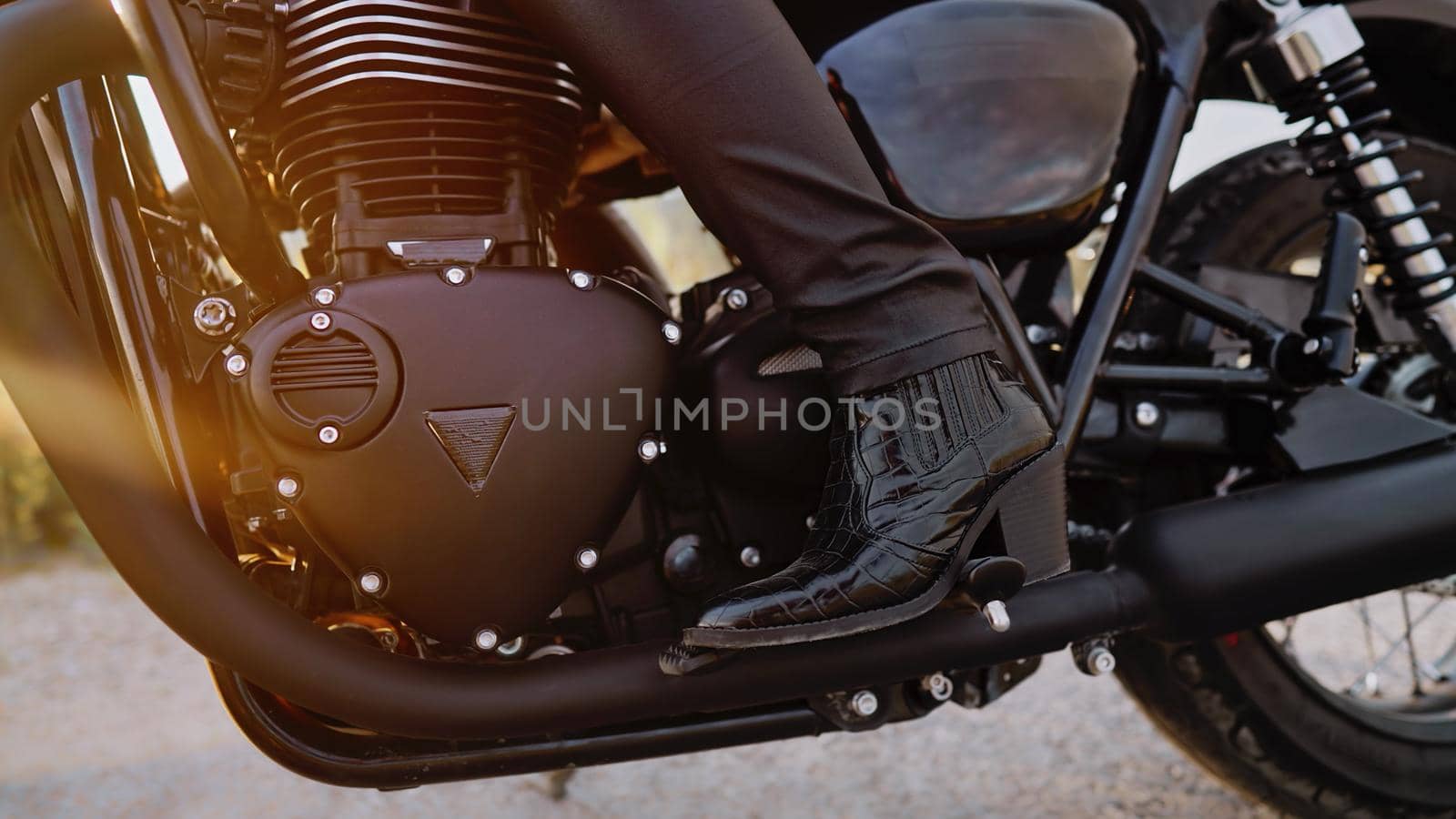 Legs of stylish motorcyclist woman sitting on classic bike. Black retro-styled motorcycle. Details of vintage design of brand-new motorbike. Sun flare, outdoor. High quality photo