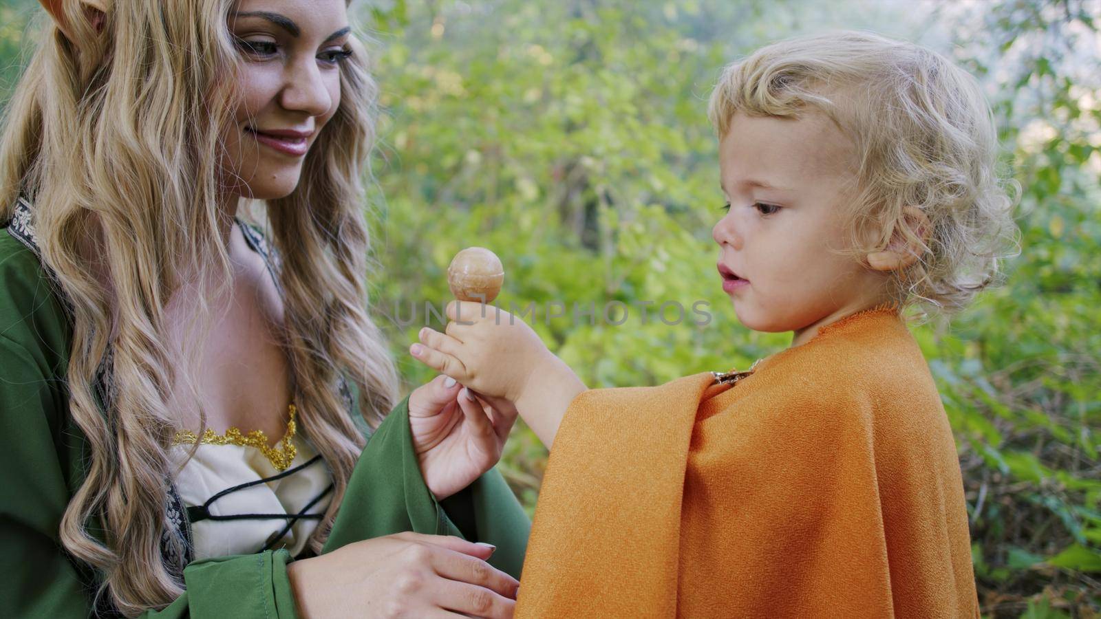 Fairy elf cosplay woman treats little baby boy hobbit with candy in green forest. Halloween concept, fairytale characters, kids. High quality photo