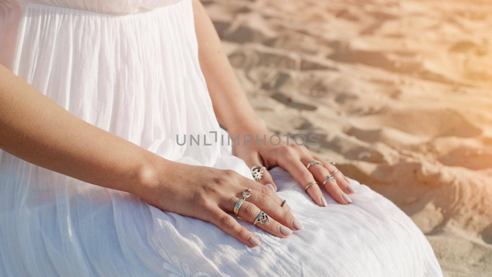 Woman in white dress and boho rings on hands sits on sandy beach alone. Meditation, immersion in inner world, unity with nature. by kristina_kokhanova
