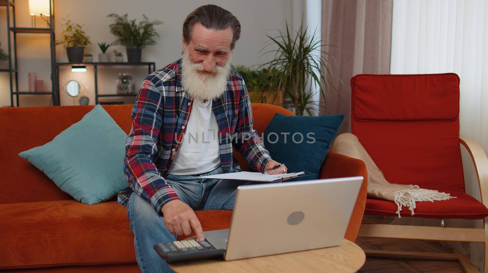 Happy smiling senior grandfather man accounting payments and calculating monthly expenses bank bills, analysing paperwork at home room couch. Elderly freelancer working online remote job on laptop