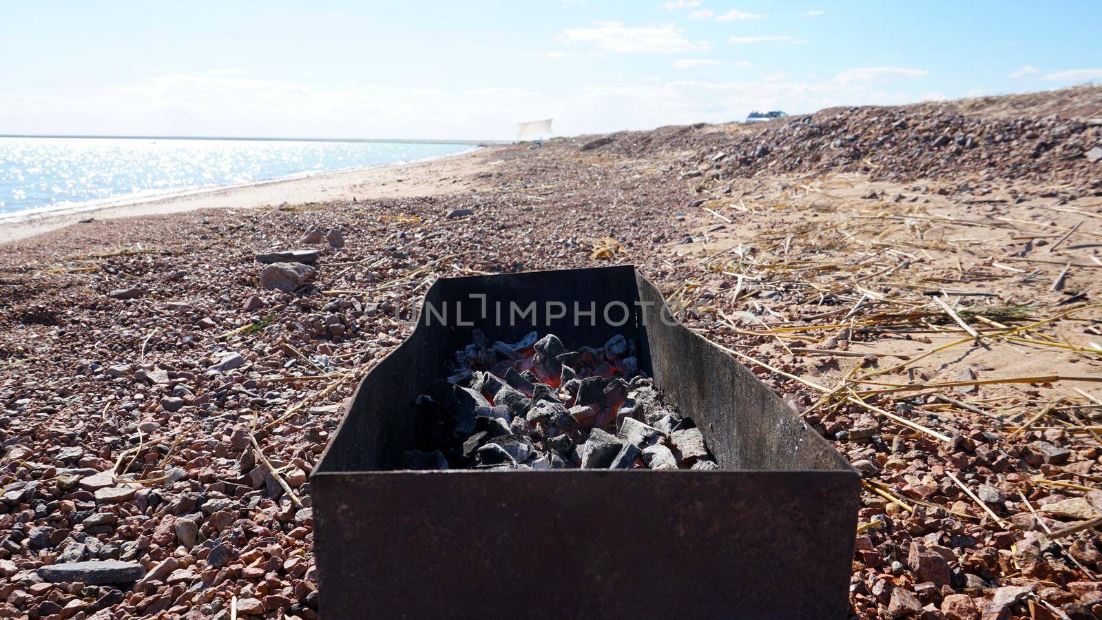 A large barbecue on the shore of the lake, there is smoke from the coals. Bright sunny day, blue lake water, yellow sand with dry branches. Preparation of coals for meat. Food in nature. Lake Balkhash