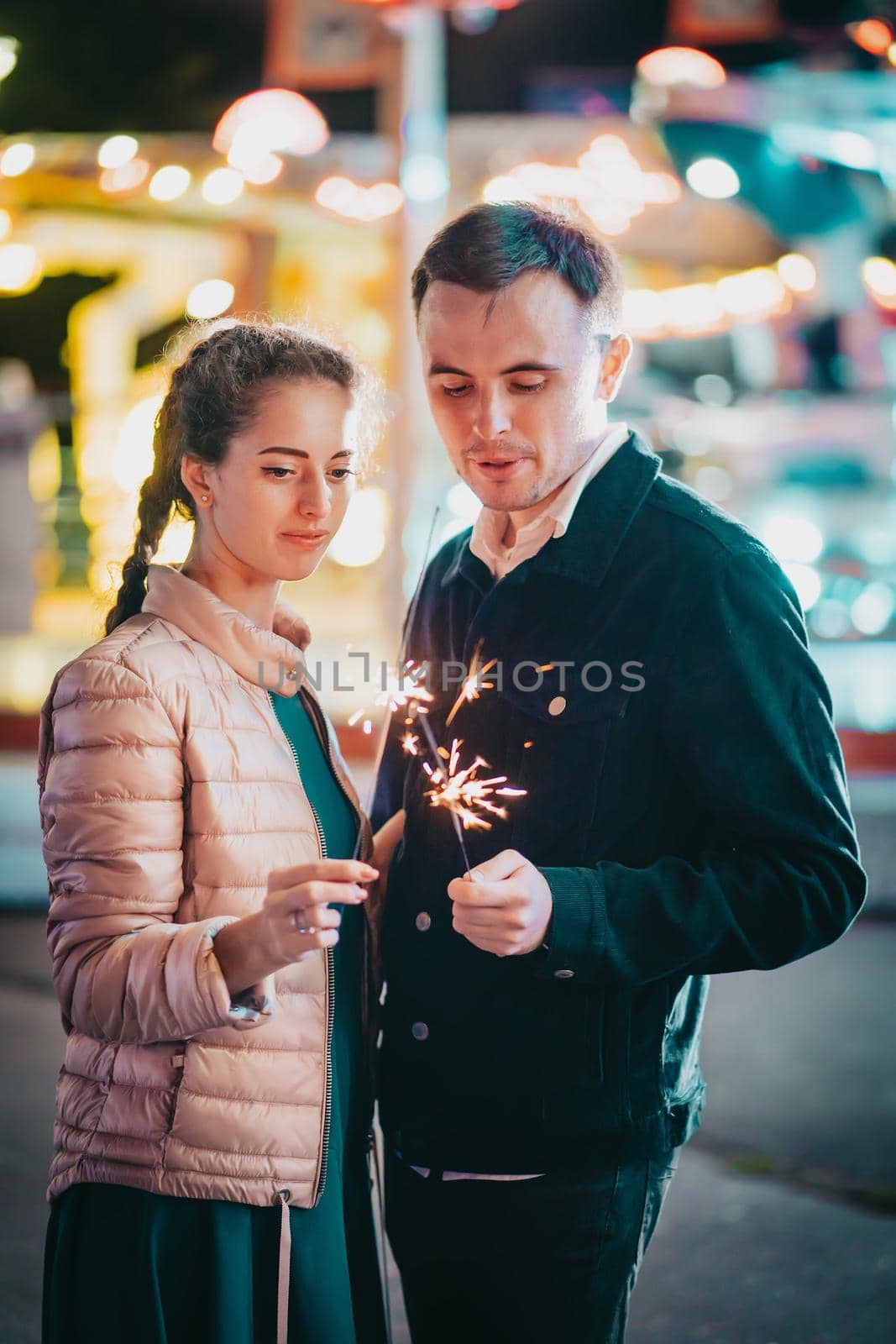 Millennial couple with sparkling bengal fire spending time together on date in amusement park at night. Lovers standing on Illuminated background. Celebration, anniversary by kristina_kokhanova