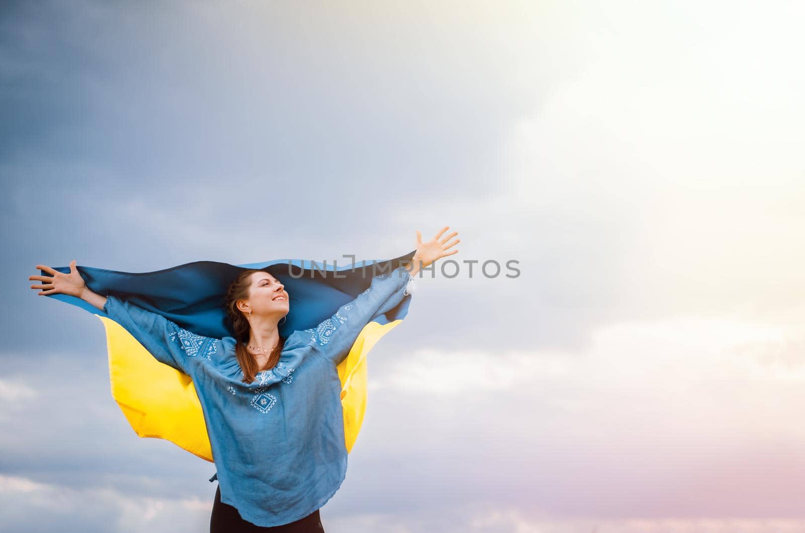 Happy free ukrainian woman with national flag on dramatic sky background. Portrait of lady in blue embroidery vyshyvanka shirt. Copy space. Ukraine, independence, patriot symbol. High quality photo
