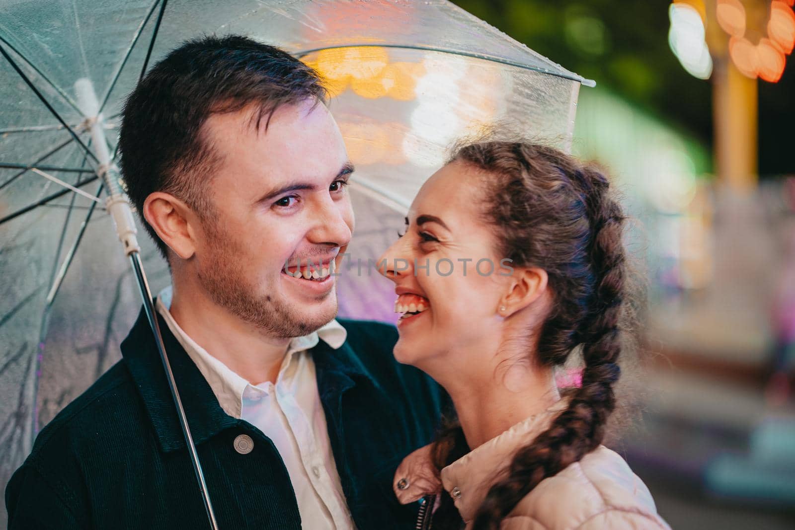 Young beautiful couple spending time together on date in amusement park at night. Rainy weather, autumn. Lovers standing under umbrella. Illuminated background. High quality photo