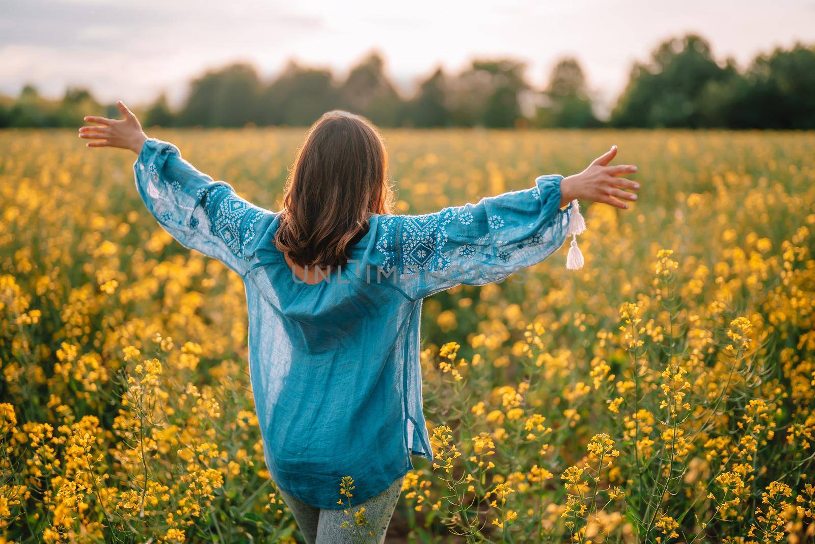 Woman standing with open arms in yellow canola field. Happy free lady in blue embroidered blouse. Alternative energy sources, oil production, biodiesel manufacturing, honey plant. High quality photo