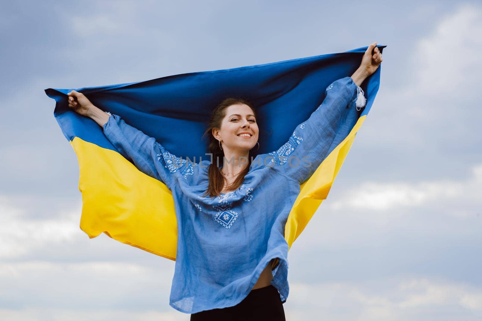 Happy free ukrainian woman with national flag on dramatic sky background. Portrait of lady in blue embroidery vyshyvanka shirt. Ukraine, independence, patriot symbol. High quality photo