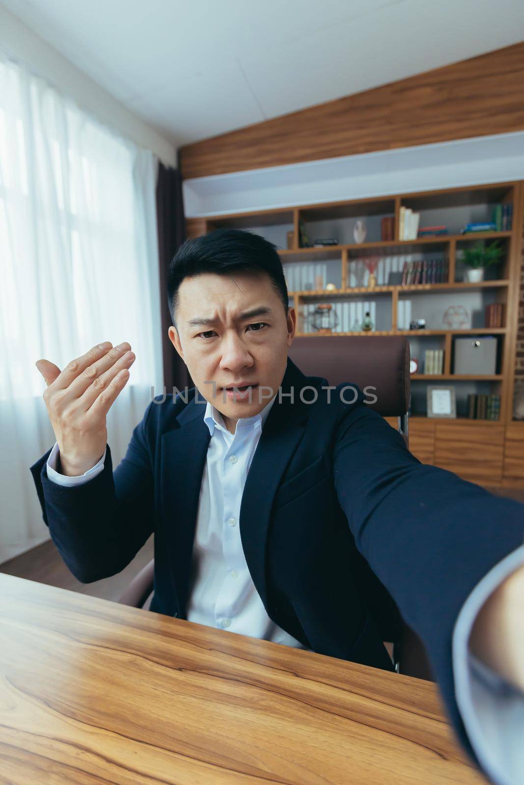 Angry Asian businessman boss talking on video call, using phone, working in classic office, arguing with employee, looking at phone camera