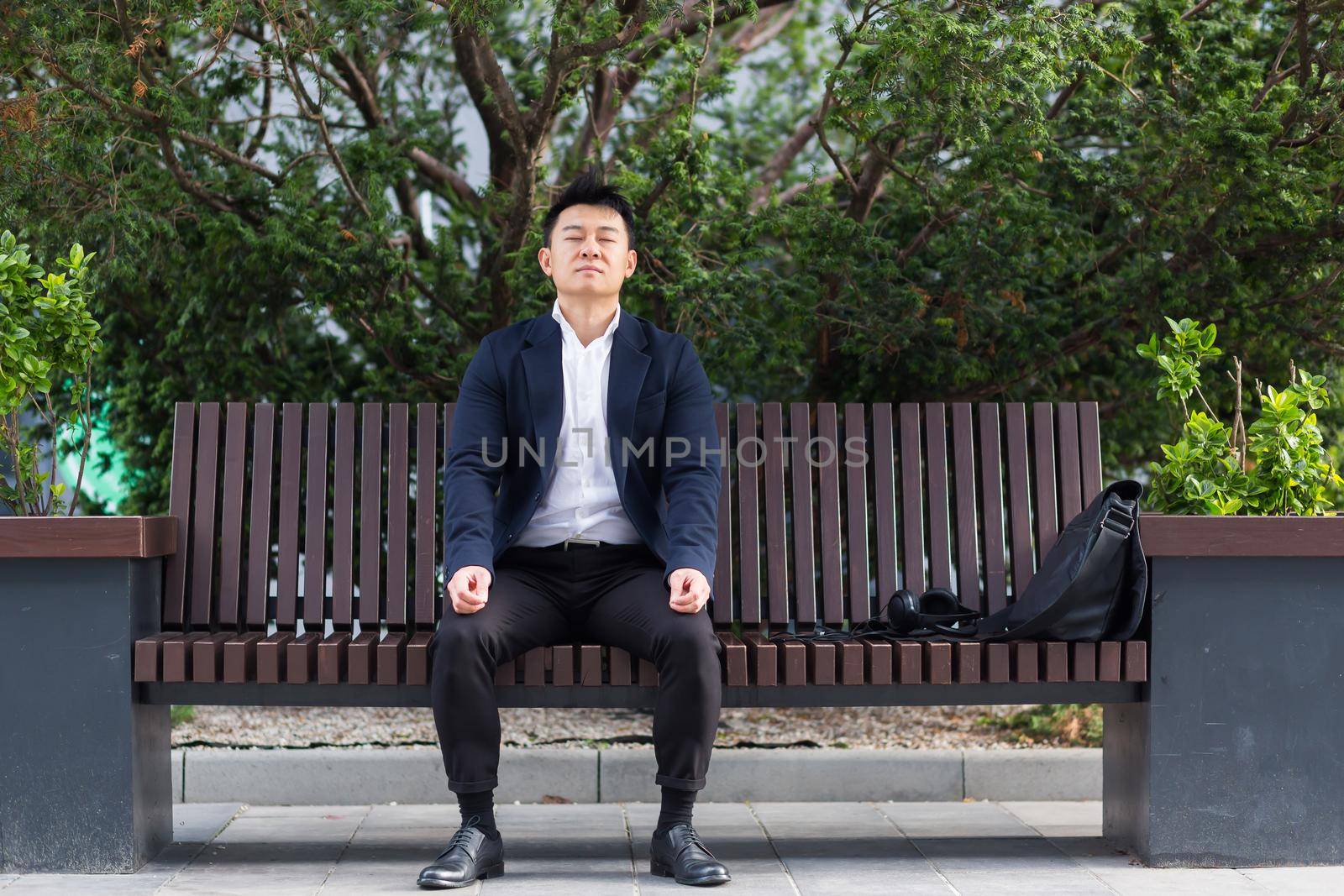 Asian businessman performing breathing exercises trying to calm stress, sitting on a bench during a lunch break in a business suit