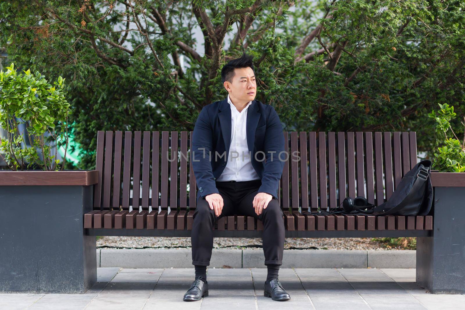 Asian businessman performing breathing exercises trying to calm stress, sitting on a bench during a lunch break in a business suit