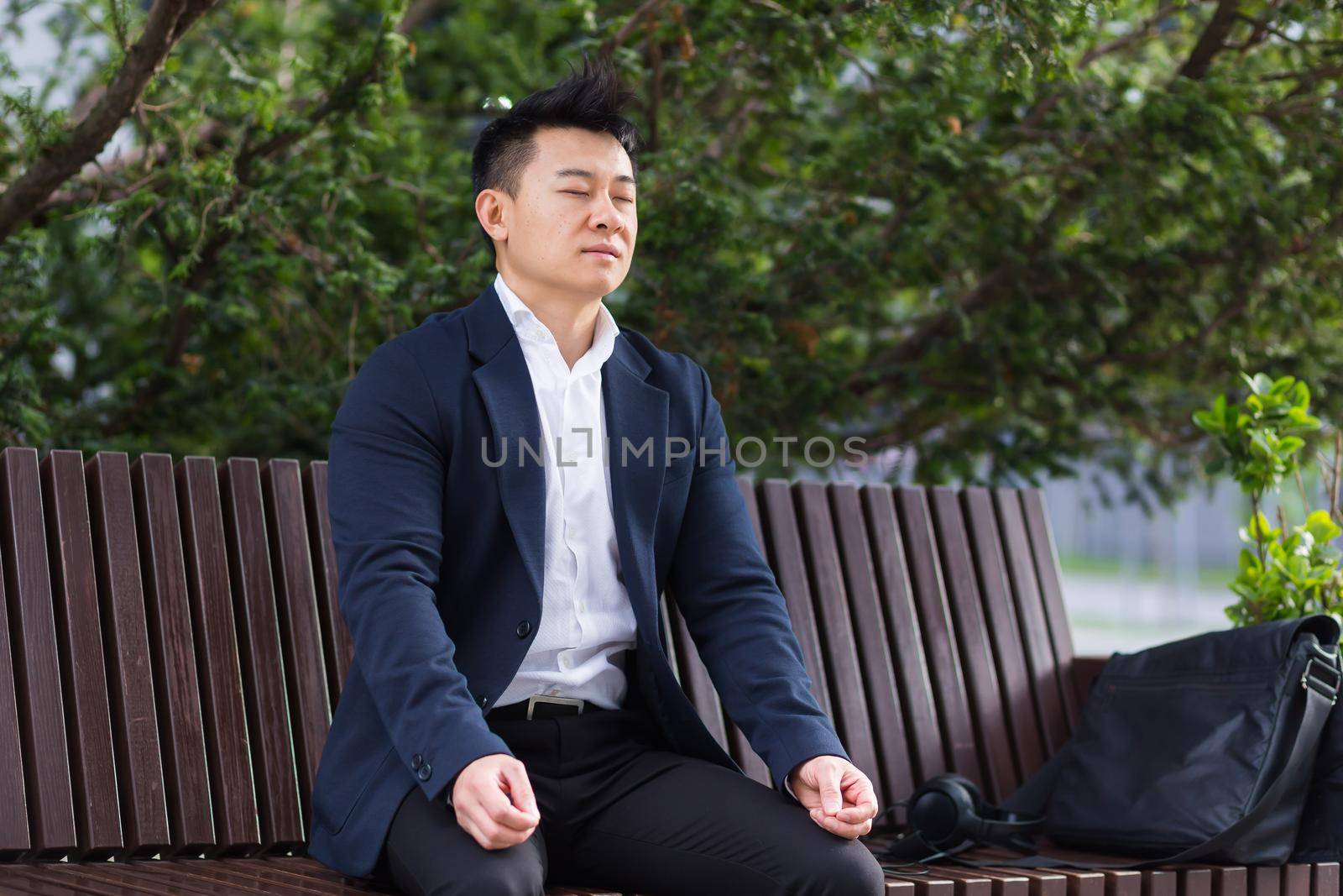 Asian businessman performing breathing exercises trying to calm stress, sitting on a bench during a lunch break in a business suit