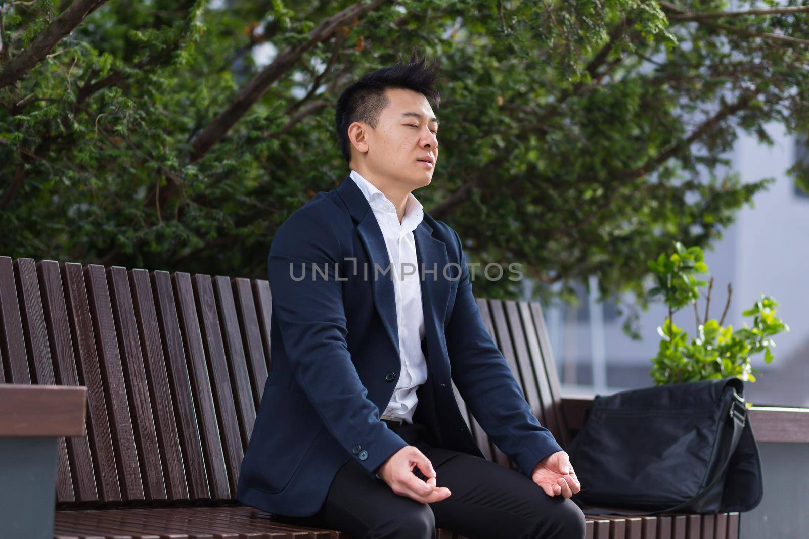 Asian businessman performing breathing exercises trying to calm stress, sitting on a bench during a lunch break in a business suit