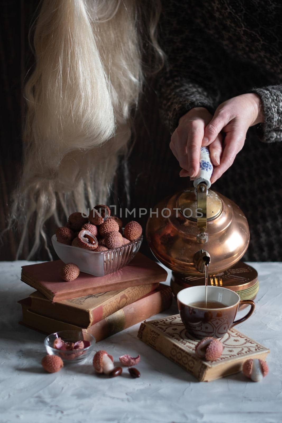A woman pours tea from a copper teapot into a cup against the background of a stack of books and a cup with lychee, homely cozy tea drinking and reading,. High quality photo