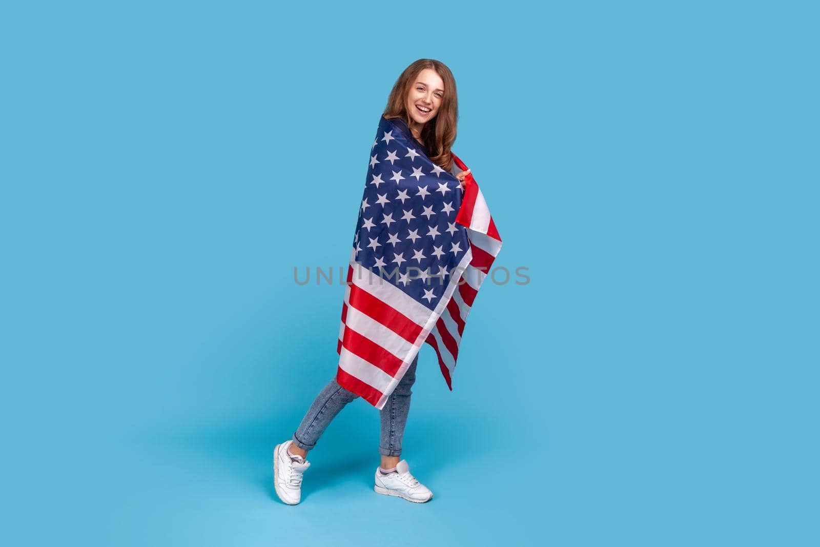 Full length portrait of smiling woman wearing striped sweater, standing wrapped in American flag, celebrating labor day, government employment support. Indoor studio shot isolated on blue background.