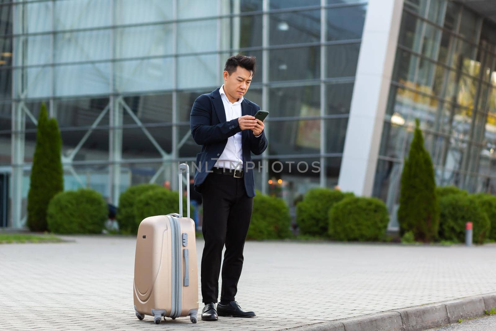 Businessman trying to call a taxi using an app and a mobile phone, asian man at the train station with a big suitcase