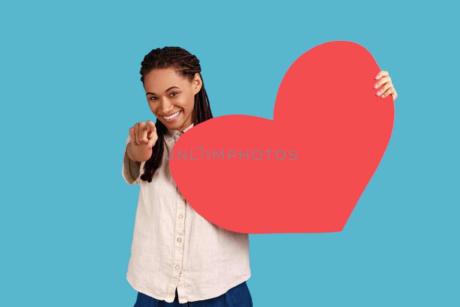 Happy cheerful woman with black dreadlocks holding in hands big red paper heart and pointing to camera, love and romance, wearing white shirt. Indoor studio shot isolated on blue background.