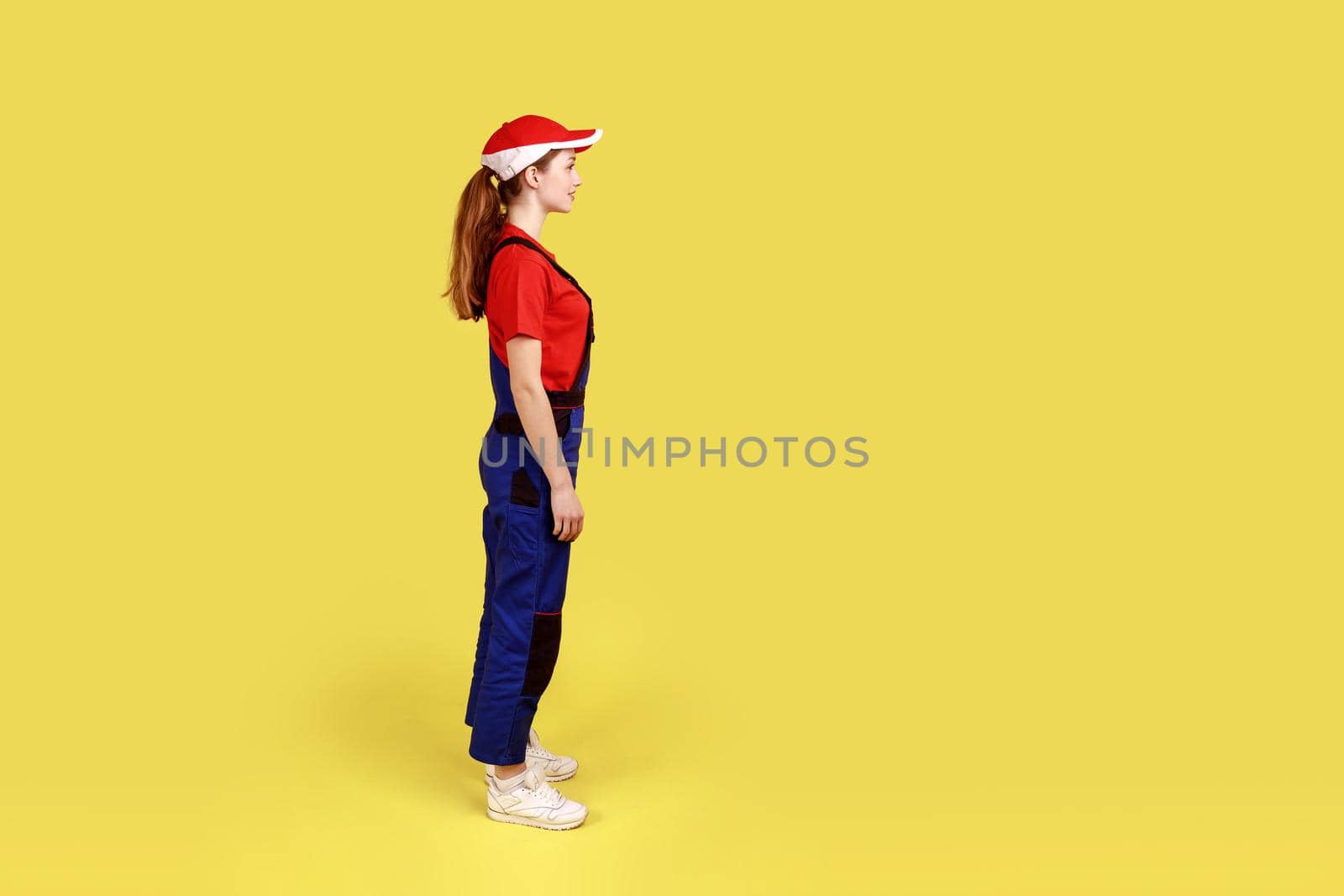 Profile portrait of worker woman standing and looking ahead with positive facial expression, being in good mood, wearing overalls and red cap. Indoor studio shot isolated on yellow background.