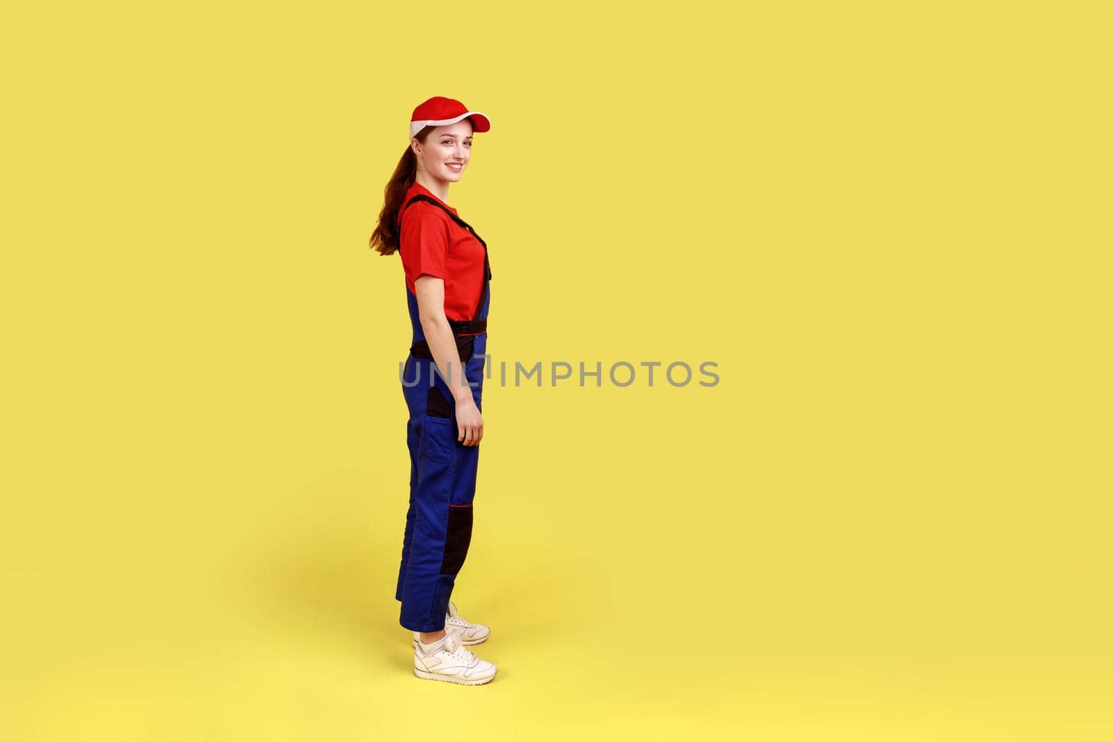 Side view portrait of worker woman standing and looking at camera with positive facial expression, being in good mood, wearing overalls and red cap. Indoor studio shot isolated on yellow background.