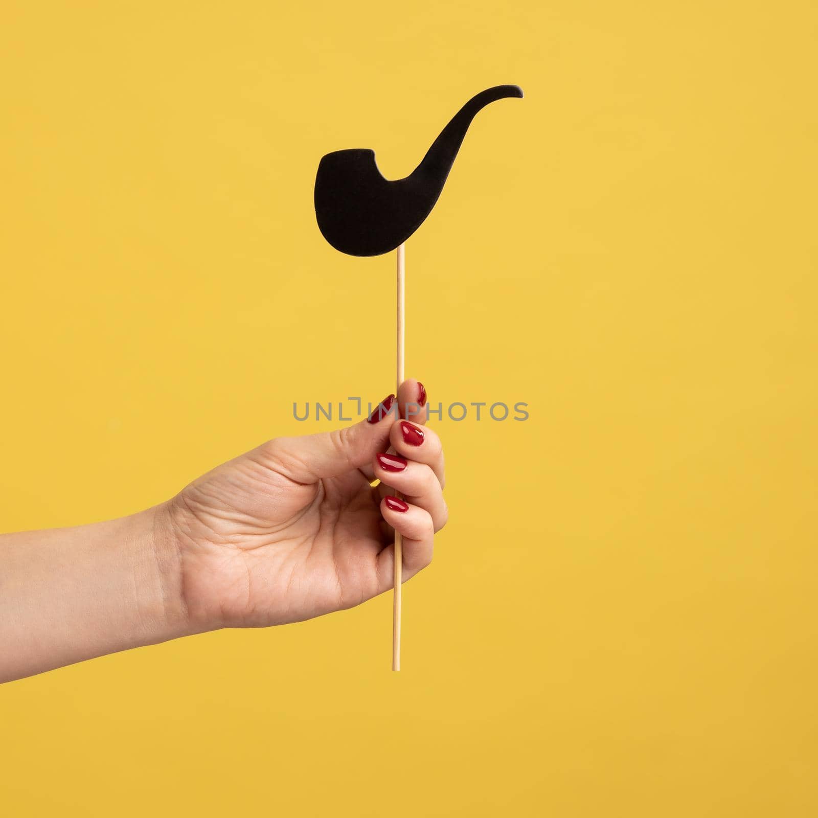 Closeup side view portrait of woman hand holding festive party props, black paper pipe on stick, celebrating holiday. Indoor studio shot isolated on yellow background.
