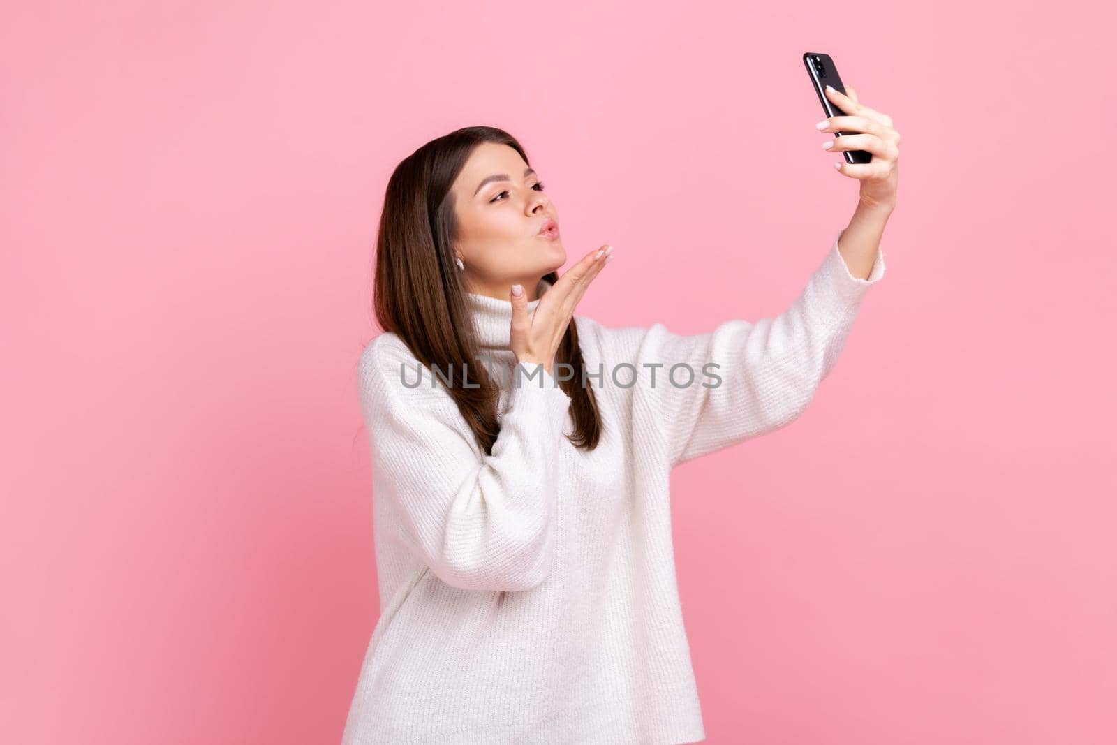 Side view portrait of romantic beautiful female sending air kisses to followers while streaming, wearing white casual style sweater. Indoor studio shot isolated on pink background.