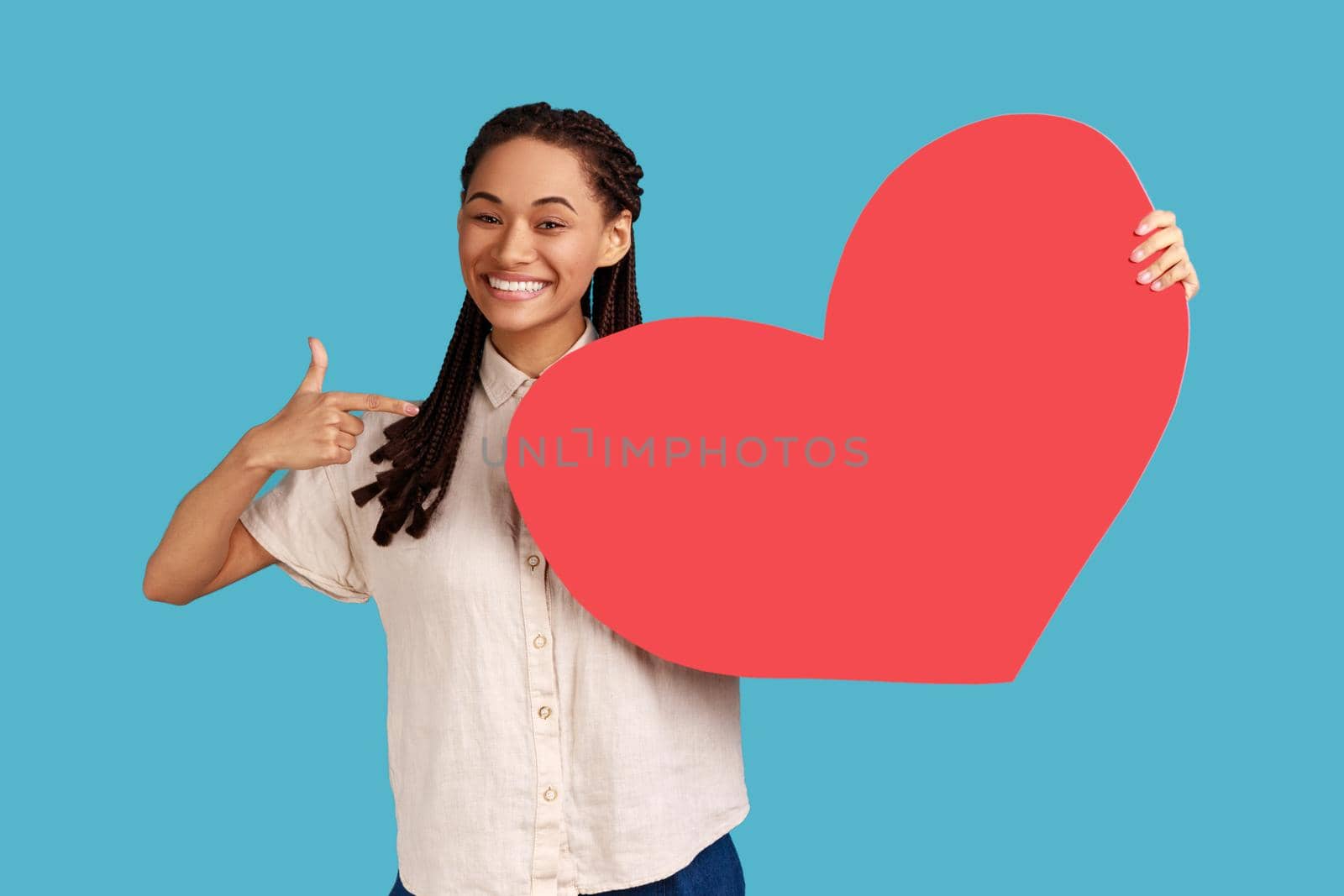 Portrait of enthusiastic woman with black dreadlocks pointing at big red paper heart and smiling, expressing extreme joy, greeting on valentines day. Indoor studio shot isolated on blue background.