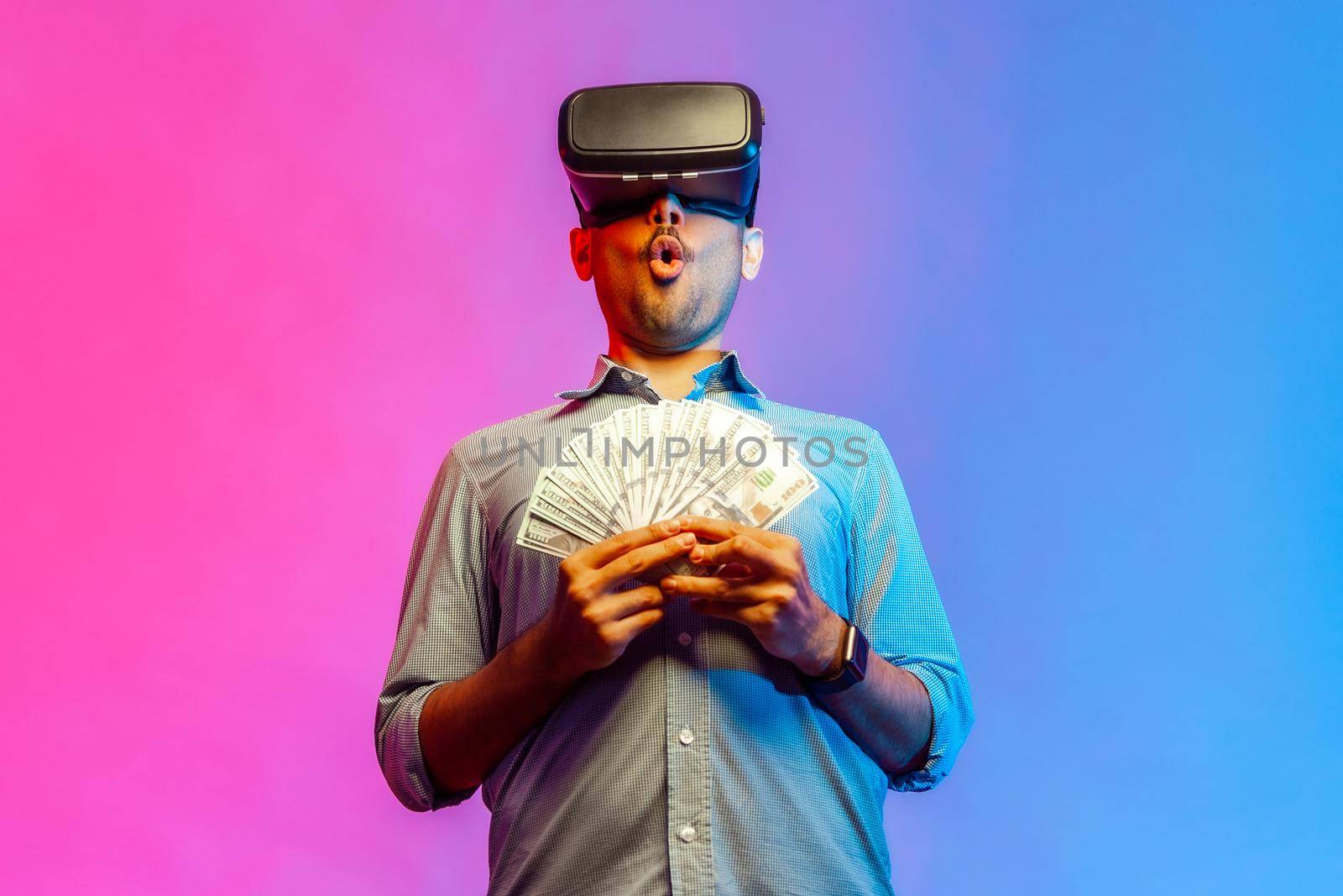 Portrait of amzed man in shirt to get money, holding dollars, illusion of rich millionaire, playing virtual reality game. Indoor studio shot isolated on colorful neon light background.