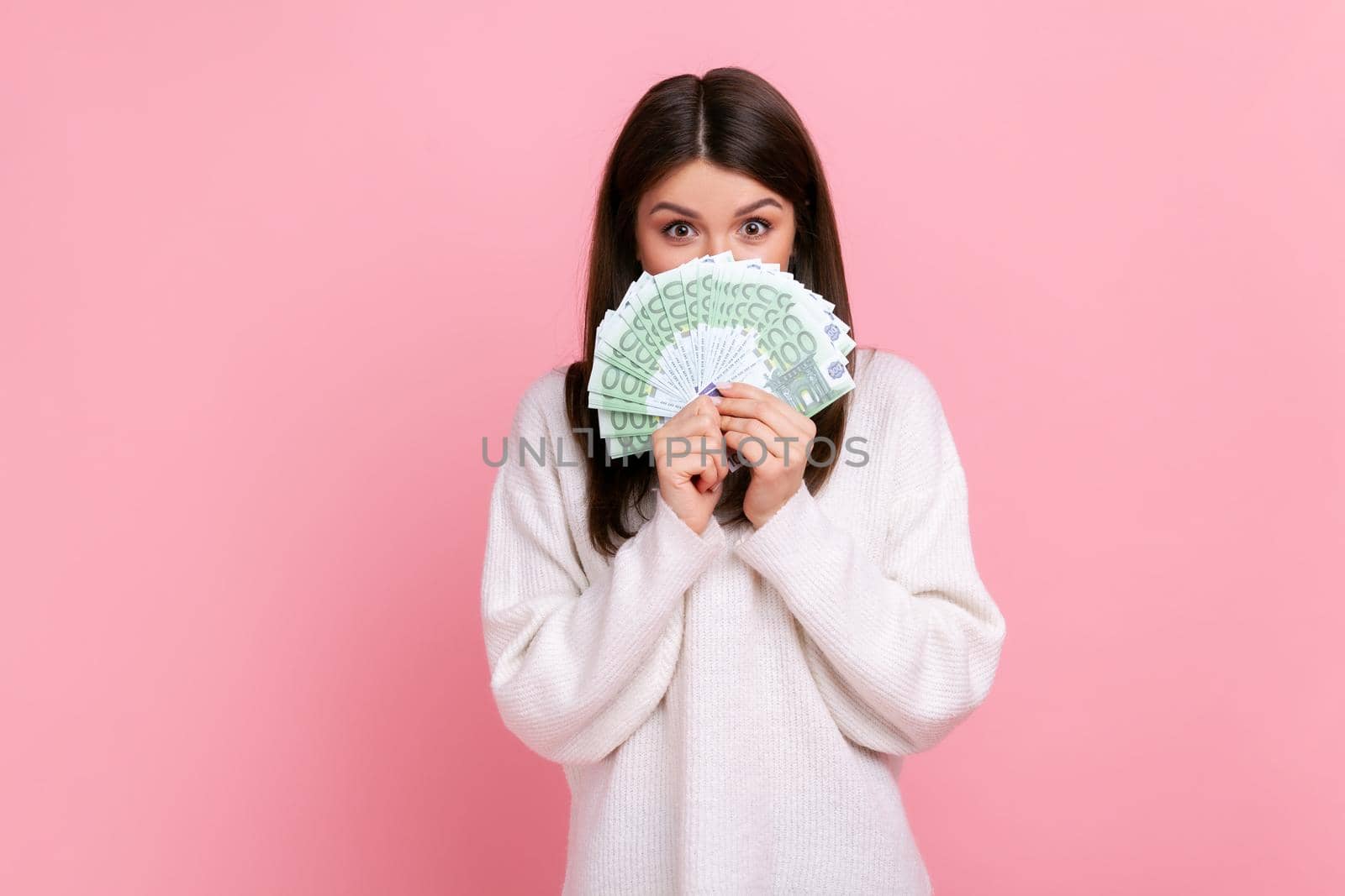 Shy brunette pretty female holding big fan of money, covering half of her mouth with euro banknotes, wearing white casual style sweater. Indoor studio shot isolated on pink background.