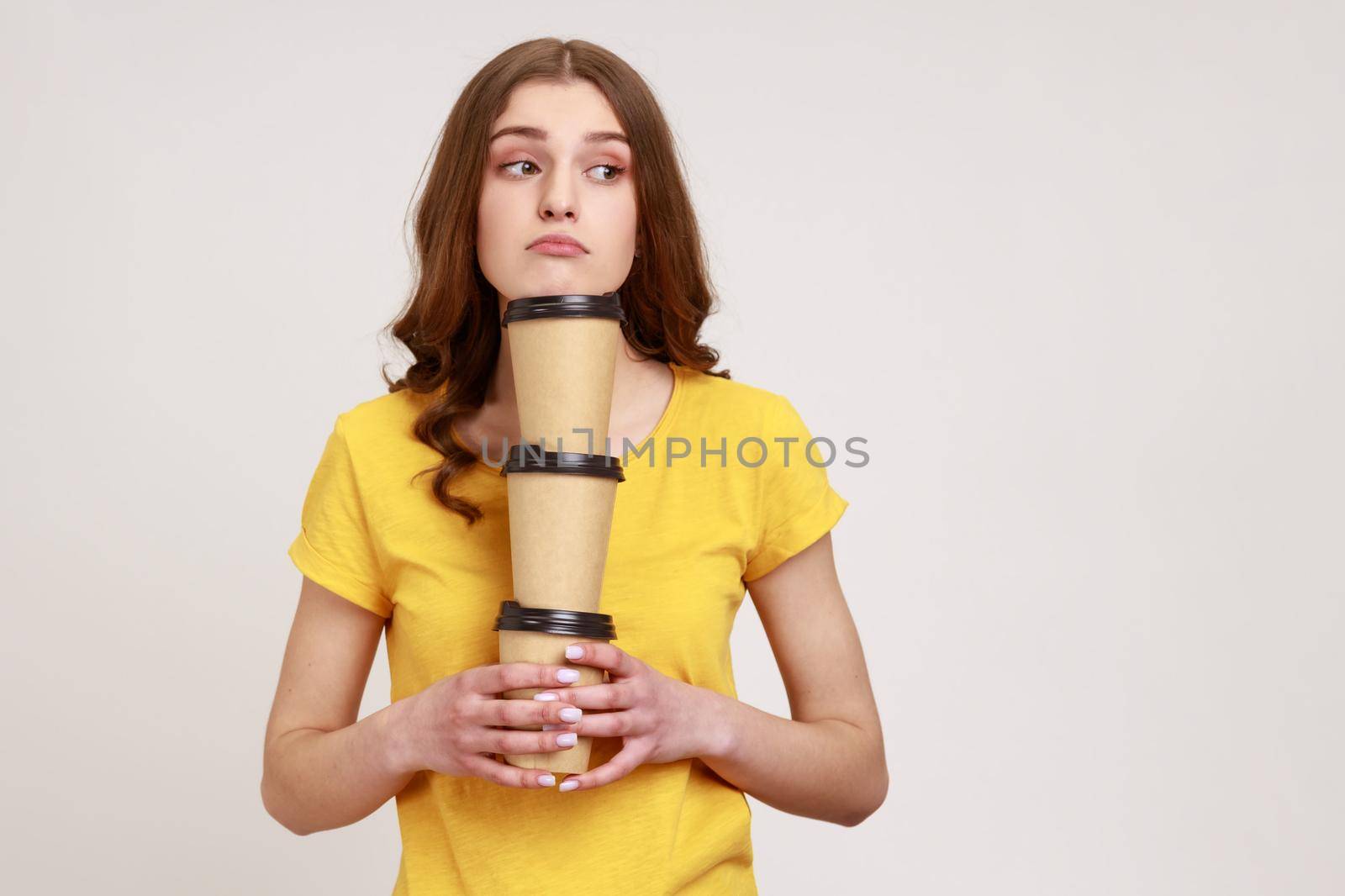 Bored sleepy teenage girl in yellow casual T-shirt holding three coffee, has exhausted expression, bringing drinks in disposable cups. Indoor studio shot isolated on gray background.