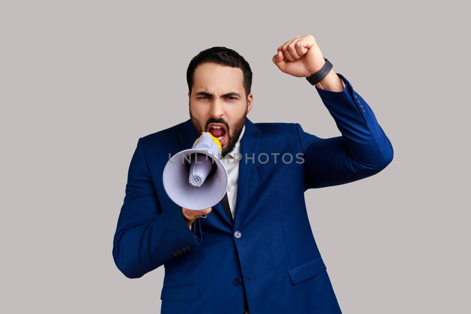 Portrait of bearded businessman standing with raised hands and holding megaphone, screaming in loud speaker, protesting, wearing official style suit. Indoor studio shot isolated on gray background.