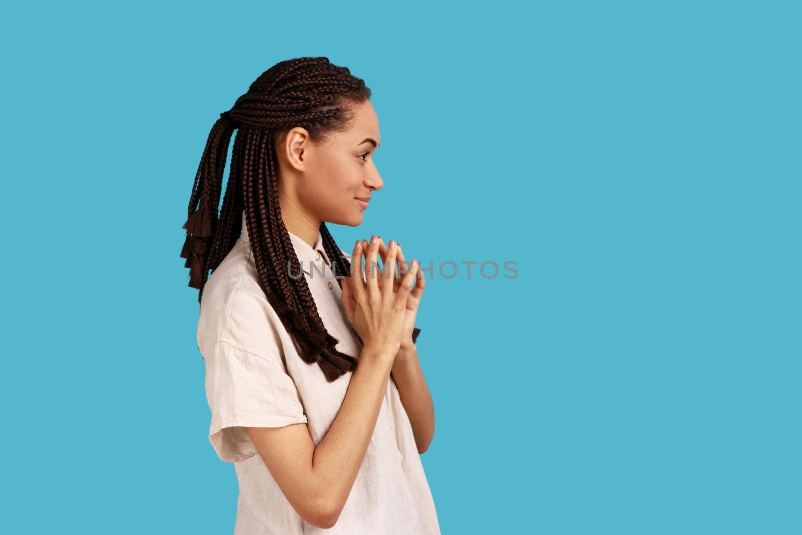 Side view of thoughtful woman with black dreadlocks schemes something, keeps fingers together, considers over cunning plans, wearing white shirt. Indoor studio shot isolated on blue background.