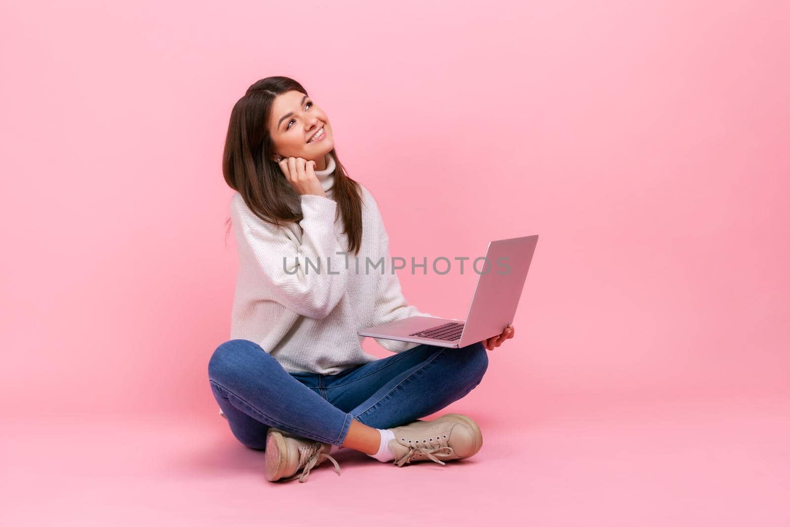 Portrait of female with dreamy facial expression, sitting with crossed legs on floor, holding laptop, wearing white casual style sweater. Indoor studio shot isolated on pink background.