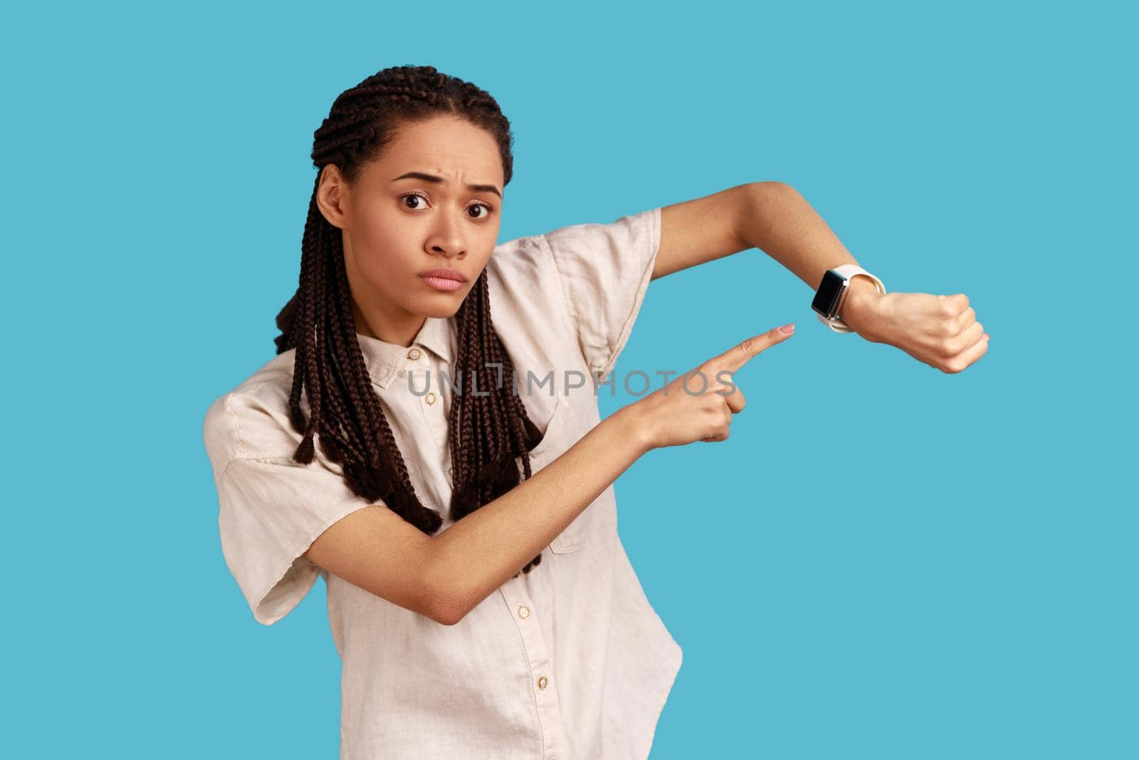 Concerned punctual woman with black dreadlocks pointing finger at smartwatch on her wrist, look at time, hurry up and act, wearing white shirt. Indoor studio shot isolated on blue background.