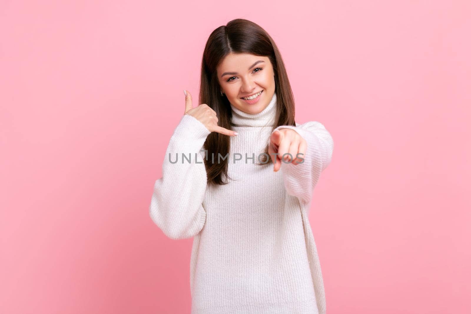 Smiling positive young adult female showing call me gesture with hand and pointing finger at camera, wearing white casual style sweater. Indoor studio shot isolated on pink background.