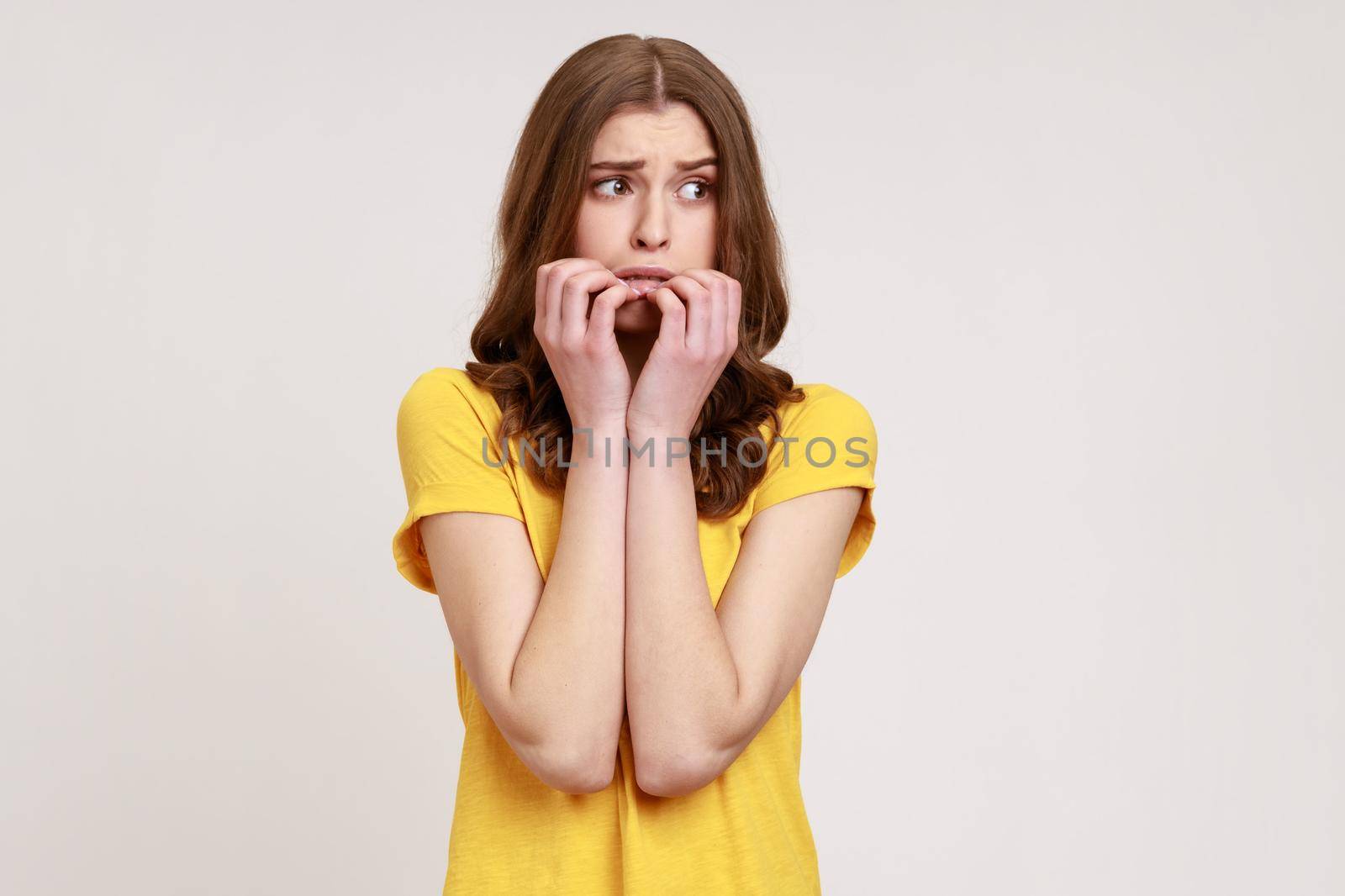 Anxious nervous female of young age with brown hair in T-shirt biting nails on fingers looking aside with terrified expression, confused and worried. Indoor studio shot isolated on gray background.